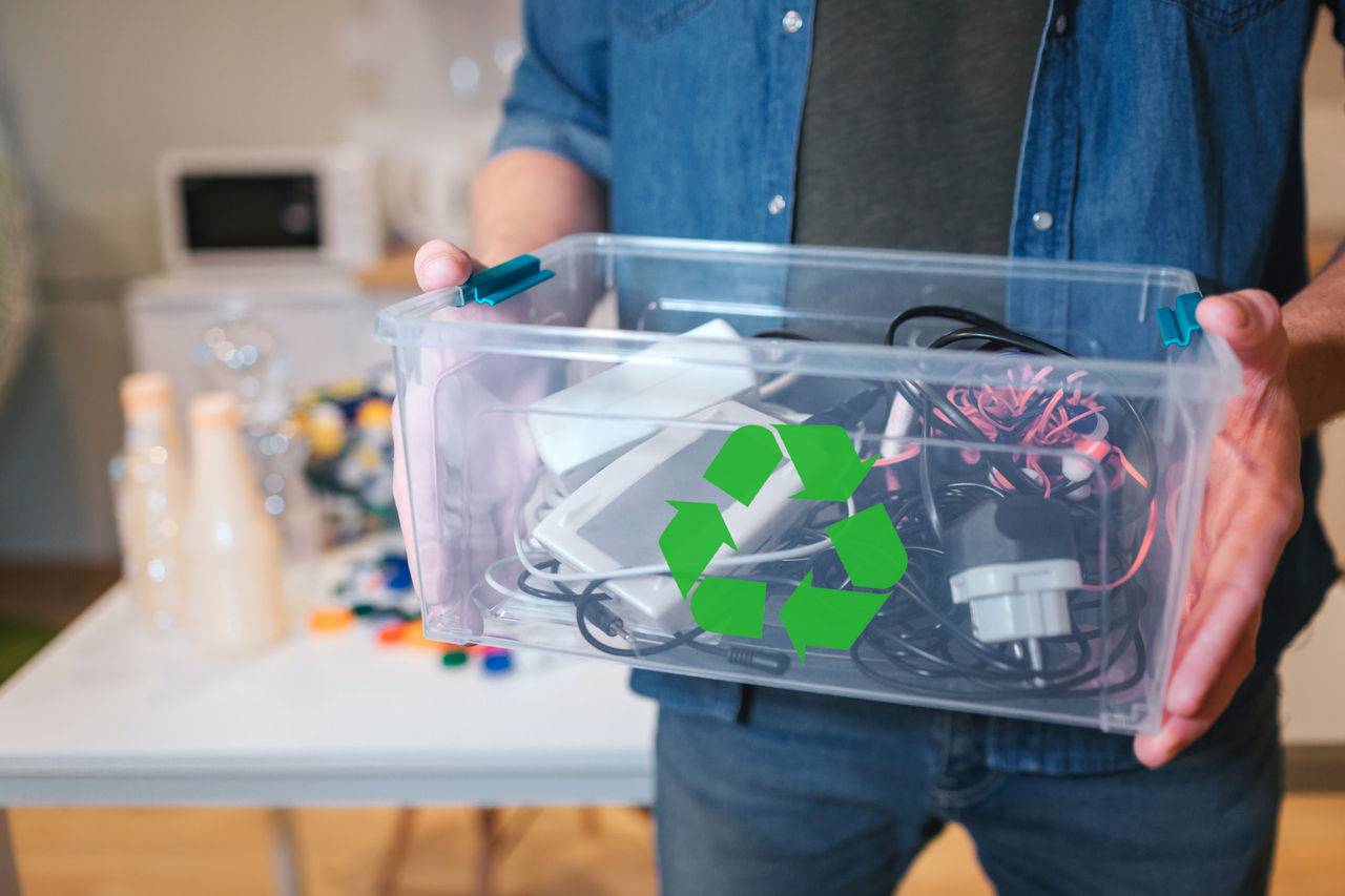 A man holds a clear box with a variety of electronic devices to recycle