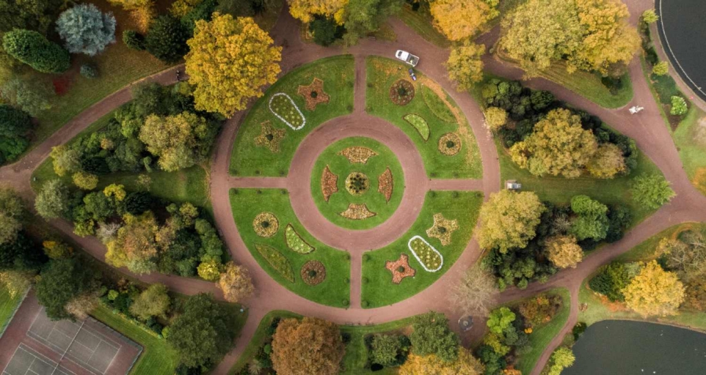 We see a circular square in a park from the sky, gardens and tree tops