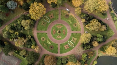 We see a circular square in a park from the sky, gardens and tree tops